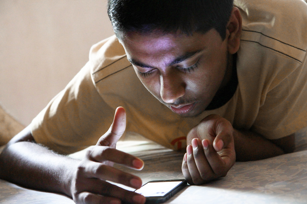A teenager holds a smartphone at a table while a parent watches from across the room, symbolizing debates over youth social media use