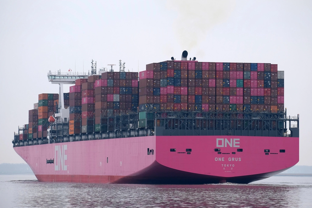 A cargo ship anchored in rough, dark waters under an overcast sky near a distant port